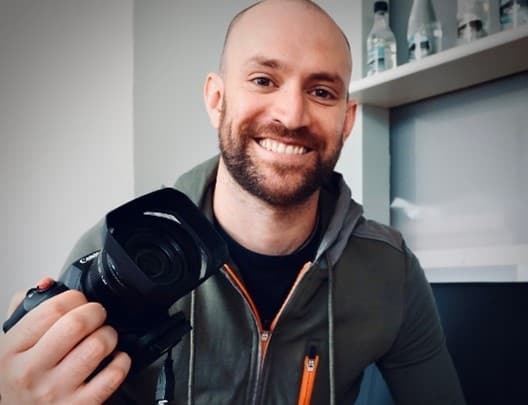 Adrian Naik, Naik Media, Bowness-on-Windermere. Adrian is wearing a hoodie and holding a camera with a smile on his face