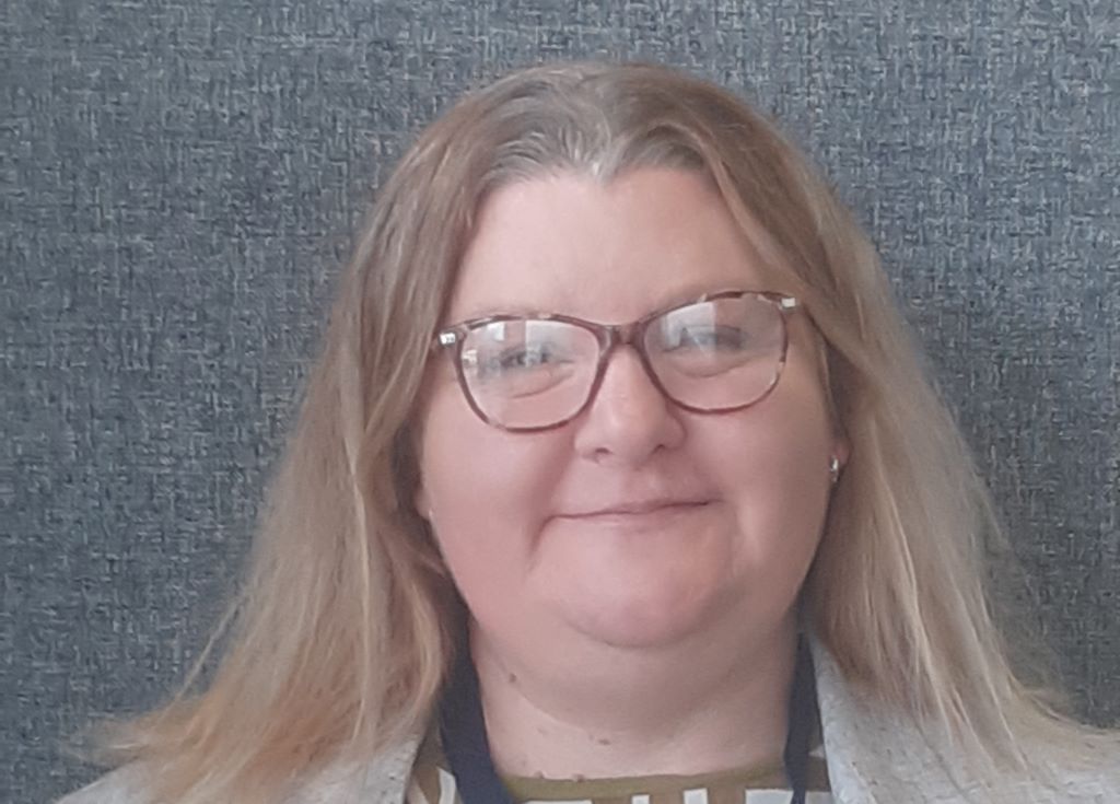 Woman smiling with glasses and blonde hair against grey background.