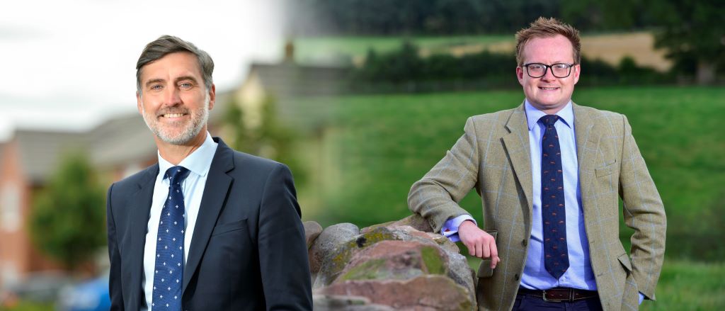 Two men in suits outdoors with fields background.